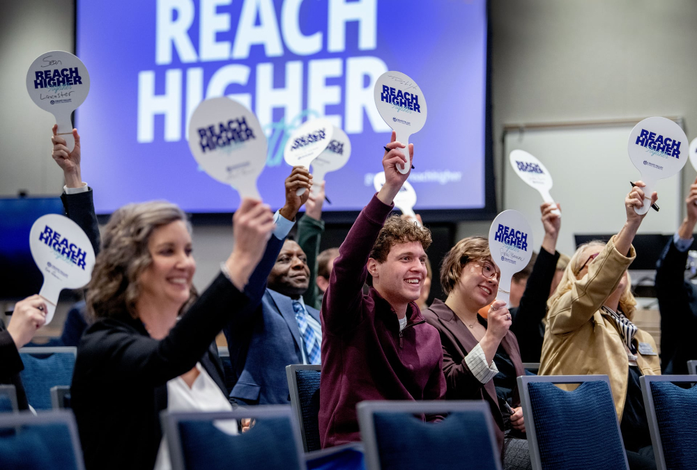 people seated in chairs holding Reach Higher paddles overhead, Reach Higher screen in background