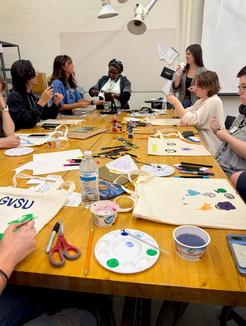 students seated around table with art supplies, painting tote bags