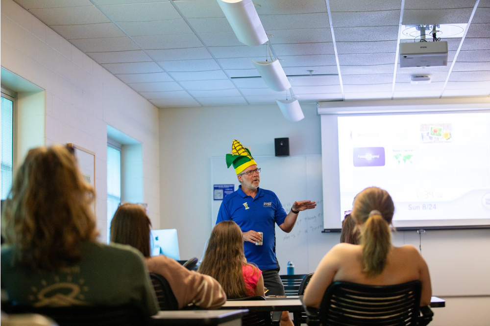 Mark Schaub in corn hat stands in front of room, students seated at tables