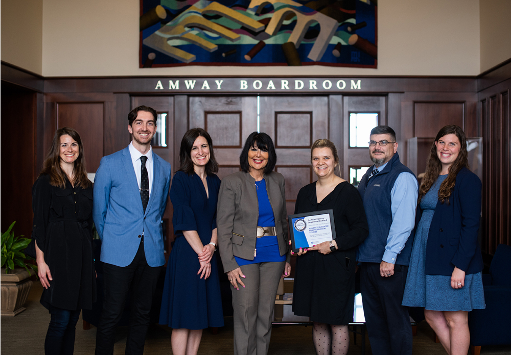 seven people standing outside heavily paneled Amway Board Room. President Mantella is in center, woman to right of Mantella holds a framed certificate.