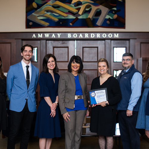 seven people standing outside heavily paneled Amway Board Room. President Mantella is in center, woman to right of Mantella holds a framed certificate.