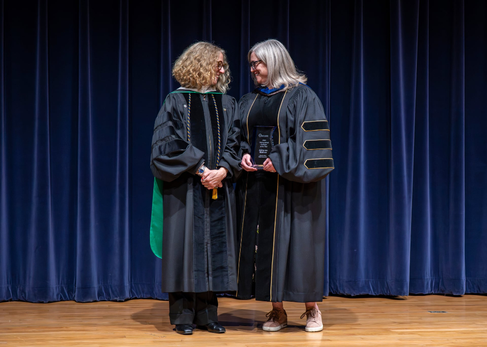 two women in academic robes on stage, blue drapery behind them