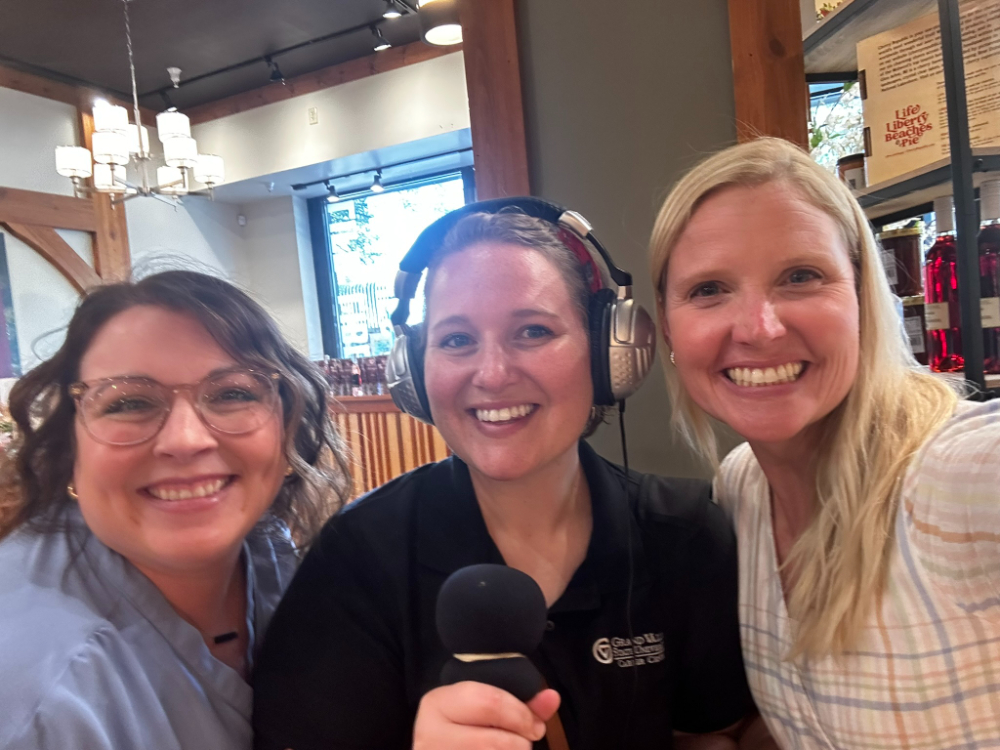three women standing in a retail store; person in middle is wearing headphones and holding a microphone for a podcast recording