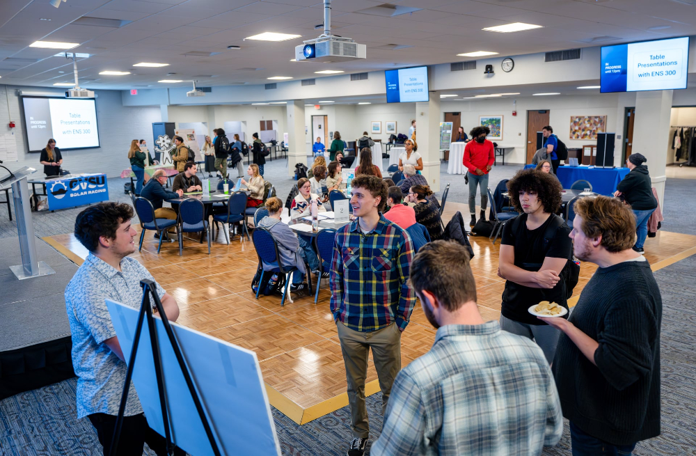 students stand around poster with people at round tables and standing in back