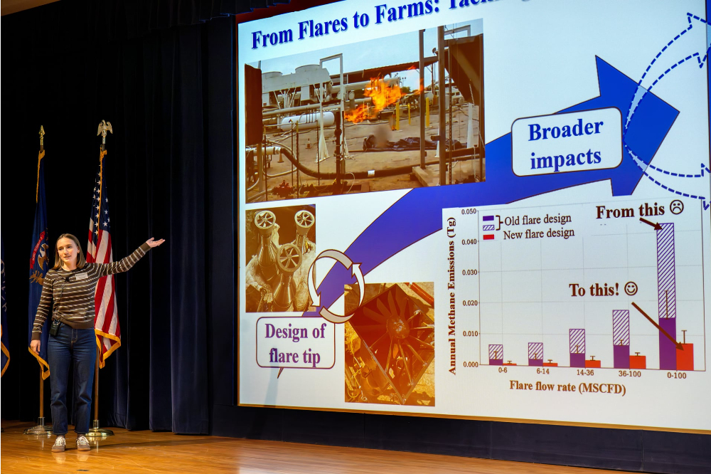 Jenna Stolzman stands on stage in Loosemore Auditorum gesturing to the screen with a slide of research