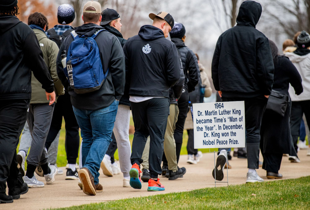 students, others walk on sidewalk during silent march for MLK event, sign reads 1964, Dr. Martin Luther King became Time's "Man of the Year" In December, Dr. King won the Nobel Peace Prize