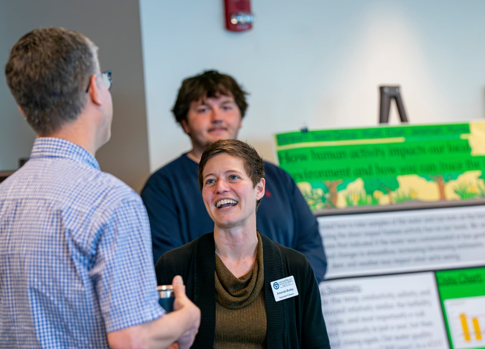Amanda Buday, center, laughs talking with someone who has his back to camera. A student stands next to a poster in the back