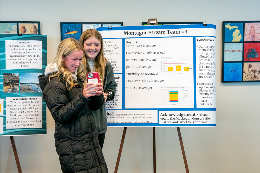 two high school students take a selfie next to a poster