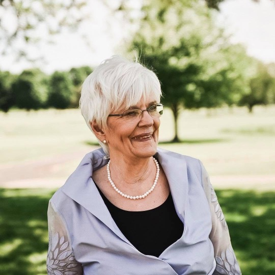 Marie Noe outdoors smiling dressed in gray jacket with dark shirt and pearls