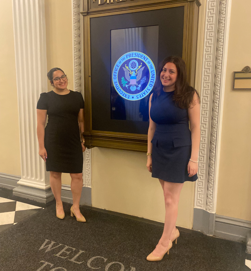 two women in dresses posing on either side of a digital directory reading the Executive Office of the President of the United States around the seal