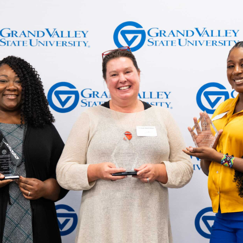 three women against a GVSU backdrop holding glass awards
