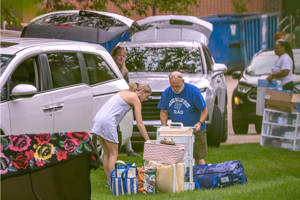 parent with Grand Valley Dad t-shirt on helps student with belongings in parking lot with SUVs with trunks open, belongings on the ground