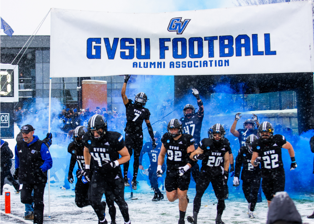 Laker football players in black uniforms running out under the GVSU football Alumni Association sign