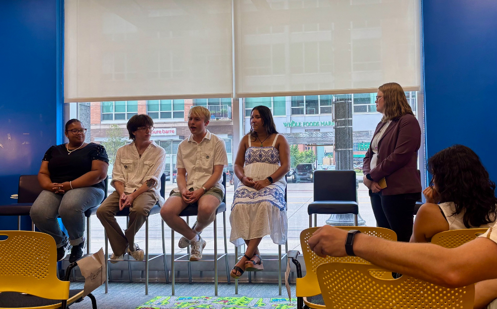 four students seated on a panel, one student stands at right