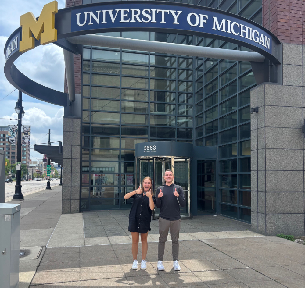 two people giving thumbs up in front of a building with University of Michigan signage