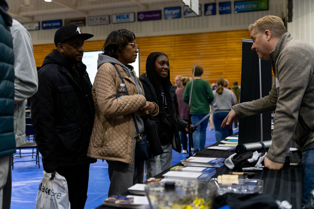 student and parents, supporters, stand on one side of a table while a man is on the other side during an open house event for prospective students in the Fieldhouse