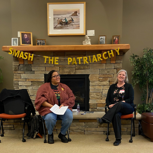two people in chairs in front of fireplace, Smash the Patriarchy banner behind them