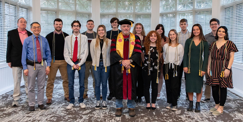 16 people standing in two rows in front of windows that look outside, person in middle is in academic regalia, others wearing honors cords