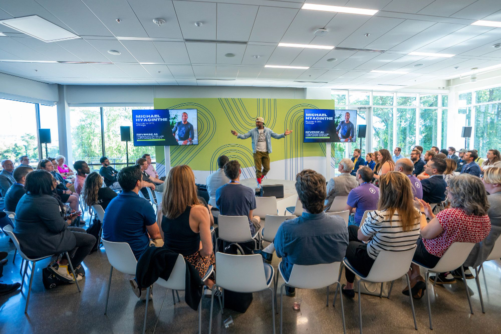 Michael Hyacinthe on stage, two screens on either side, background of green with blue swirls to represent Tech Week Grand Rapids