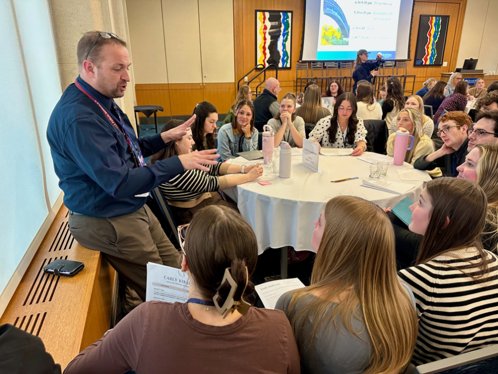 man leaning against wall before round table of students holding paperwork