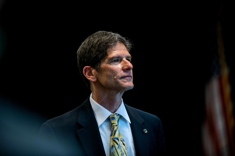 portrait of Alan Steinman, who is standing near an American flag in a blue suit jacket and colorful tie