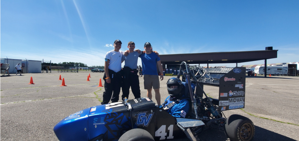 mini Formula One car on racetrack with driver in helmet and three students standing behind it