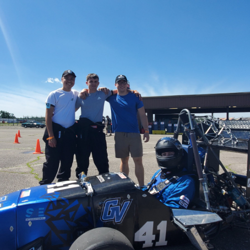 mini Formula One car on racetrack with driver in helmet and three students standing behind it