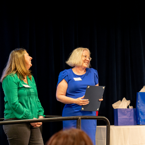 two women laugh as they stand on stage; Chelsea Ridge in green and Kathy Agee in blue dress