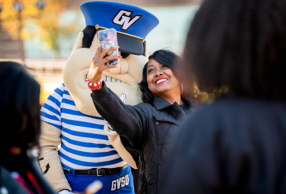 graduate in black coat takes a selfie with Louie the Laker