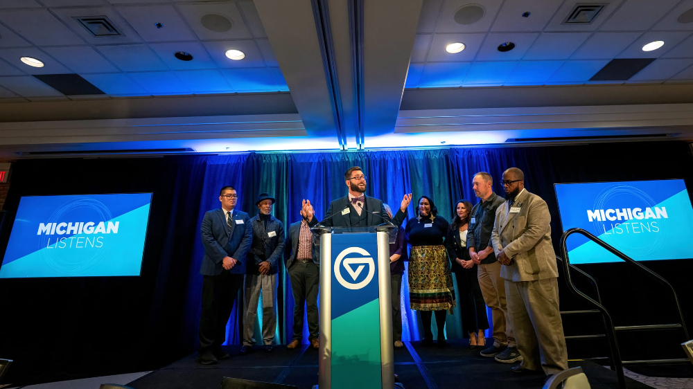 Kyle Kooyers stands at podium with nine people in semicircle behind him