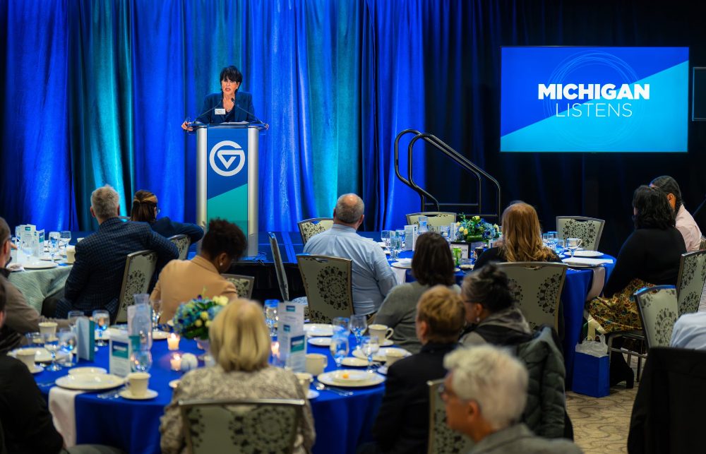 President Mantella at podium, people seated at round tables, Michigan Listens on screen at right