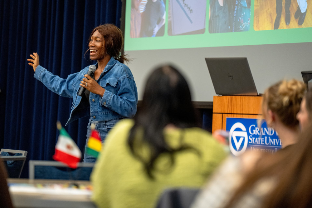 student in denim jacket with arm raised, speaking in front, people at round tables