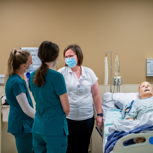 a high-fidelity manikin is in a hospital bed while two nurses in green scrubs and a woman in street clothes talk on the left side of the bed