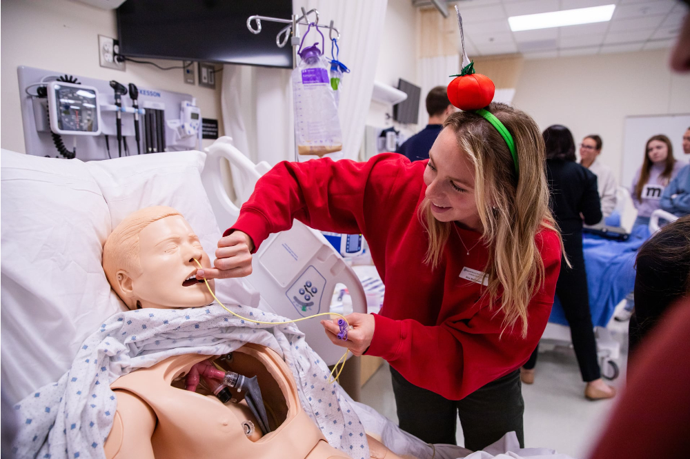 A health professions student in red sweatshirt and apple hairband practices nasogastric tube placement on a simulation model.