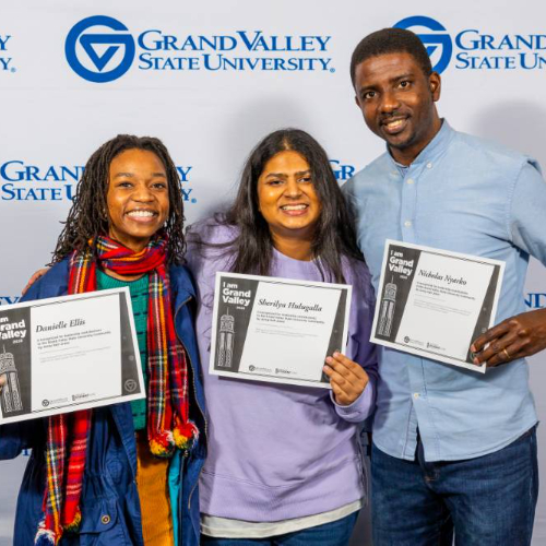 three students hold certificates in front of a background of the GVSU logo