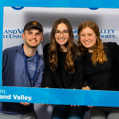 three students hold a photo prop frame with "I am Grand Valley" in the lower left corner