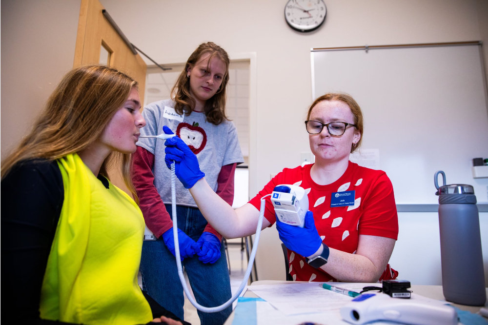 one student in red tshirt takes the temperature of another student in banana costume using an oral thermometer. third student stands in background