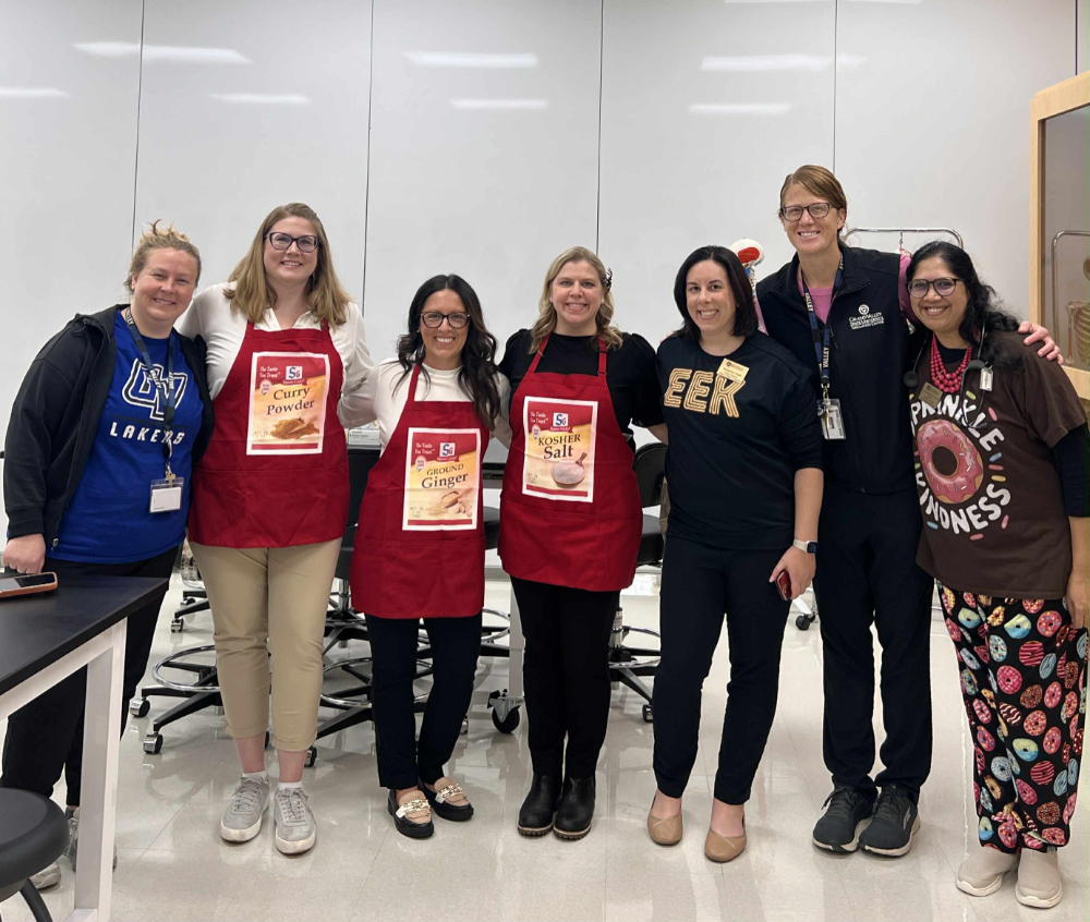 seven people standing with arms around each other, some in costumes like an apron or donut sweatpants