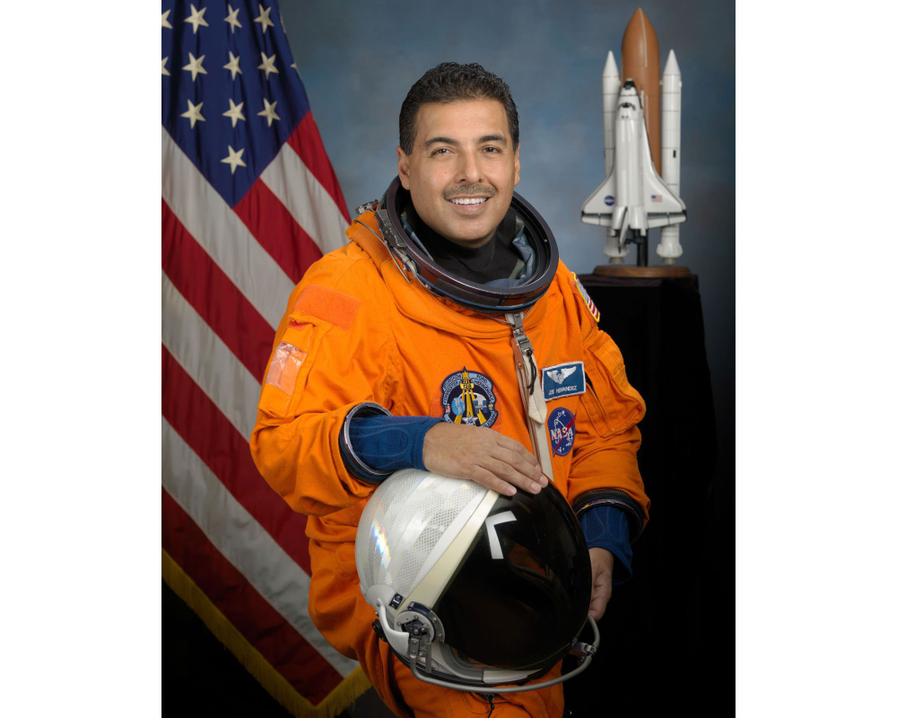 NASA astronaut José Hernández is in front of a backdrop of an American flag and spaceship; he is in an orange uniform and holding a helmet