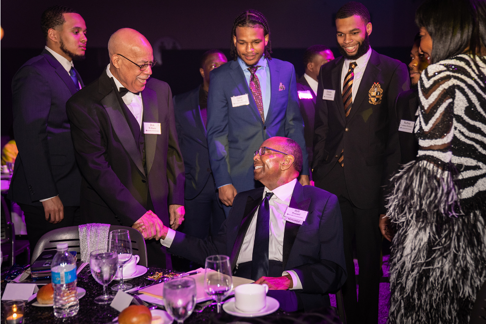 man seated at table, four people in tuxes or suits stand behind to talk