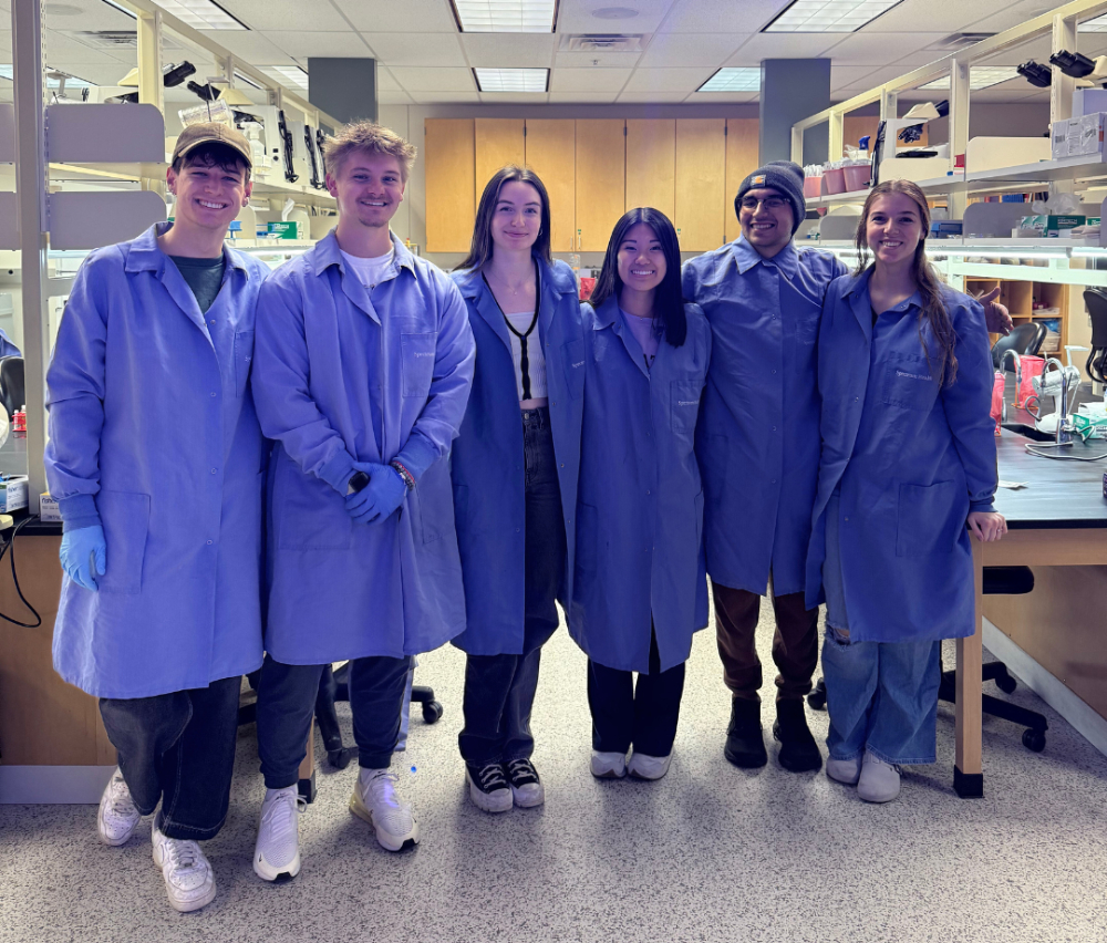 six students in blue, long lab coats standing in a lab