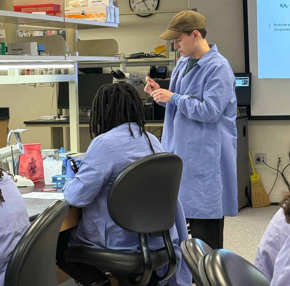 GVSU student Luke Markley stands at a lab station in long blue lab coat, helping a KCTC student who is seated