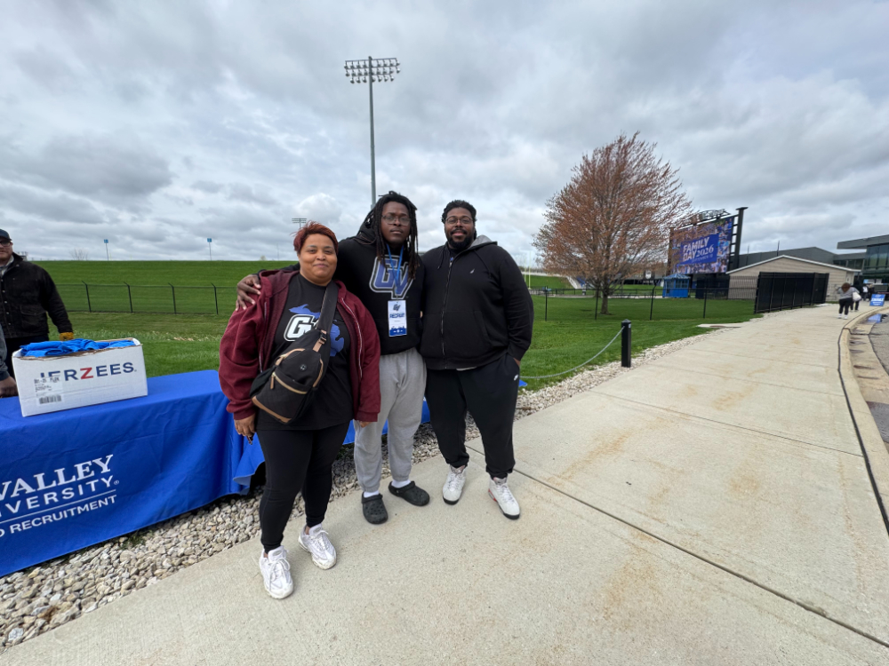 three people standing near table with blue GVSU draped tablecloth, person in middle has lanyard on with large tag, Recruit