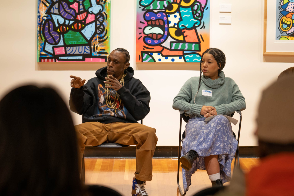 two people sitting in chairs answering questions from audience in art gallery; brightly colored artwork is on the walls behind them