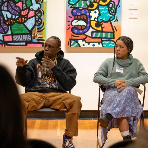 two people sitting in chairs answering questions from audience in art gallery; brightly colored artwork is on the walls behind them