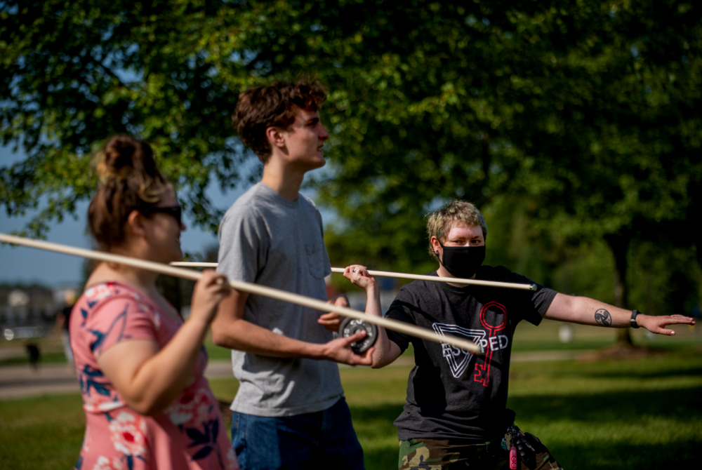 three students stand outside and hold wooden spears