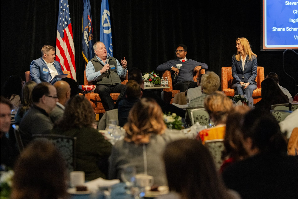 four people seated in chairs on stage at the Eberhard Center; US flag, Michigan flag and GVSU flag are behind them. People seated at tables in front