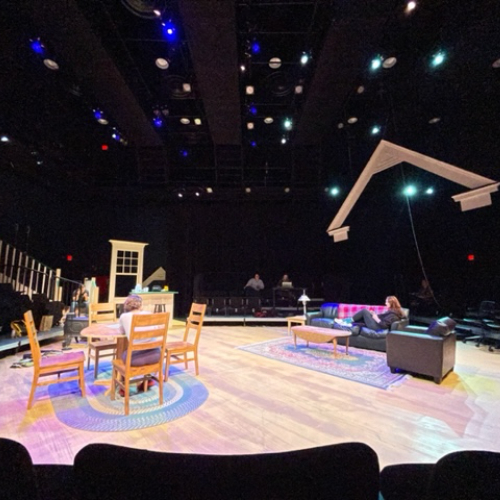 a stage during rehearsal with a piece of roof suspended on wire on the right, table and chairs on the left