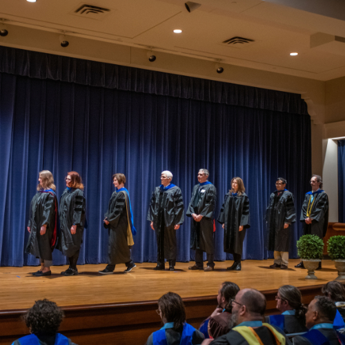 eight faculty members in academic regalia lined up on stage, blue curtain behind them