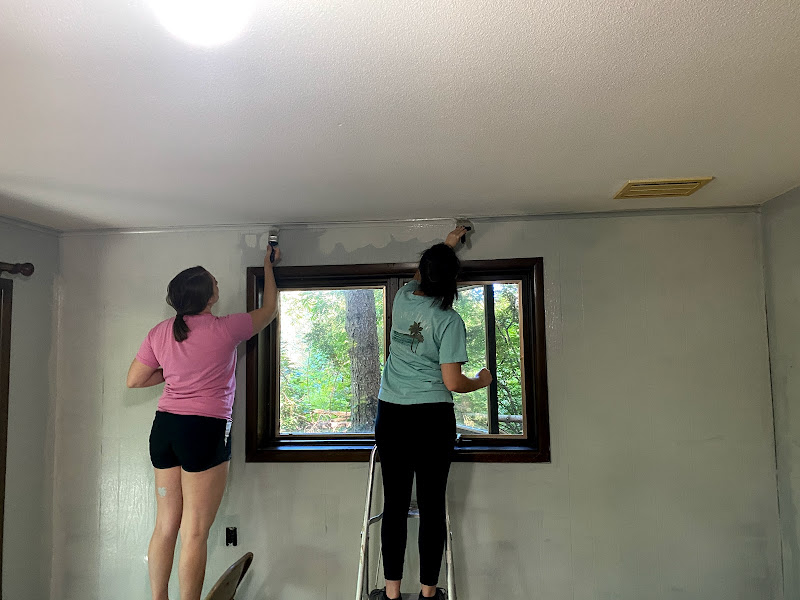 two students, standing on a chair and short ladder, paint a wall near a ceiling of a home